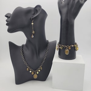 Pode incluir: Um conjunto de joias douradas que inclui um colar, uma pulseira e brincos. O colar e a pulseira têm um design de elos de corrente com pequenas contas redondas iridescentes e um amuleto oval maior. Os brincos são brincos pendentes com um design de elos de corrente e pequenas contas redondas iridescentes.