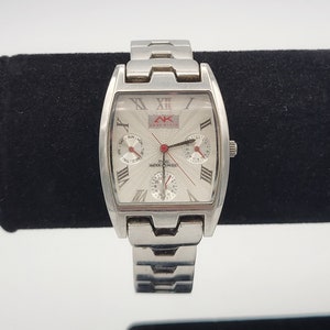 Puede incluir: Un reloj de pulsera plateado con una esfera rectangular. El reloj tiene una correa plateada y una esfera blanca con marcadores de hora en números romanos negros. La esfera del reloj también presenta tres esferas más pequeñas, una ventana de fecha y el texto "AK Anne Klein" en rojo.