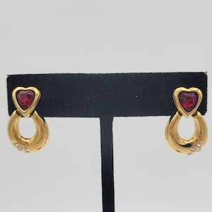 Puede incluir: Pendientes de tono dorado con una piedra roja en forma de corazón y detalles de cristal transparente.