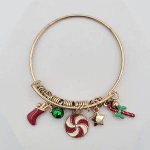 May include: Gold-tone Christmas charm bracelet featuring festive charms. Charms include a red stocking, green ball, red and white candy, gold star, and a red and green candy cane. The bracelet is a bangle style.