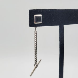 Puede incluir: Un broche de corbata plateado con una piedra cuadrada negra y una cadena.