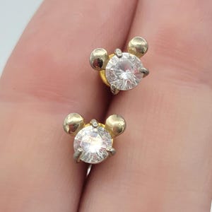 Puede incluir: Un par de pendientes de bot&oacute;n dorados. Cada pendiente presenta una piedra clara, redonda y brillante en el centro, rodeada por un marco dorado con dos peque&ntilde;as esferas doradas en la parte superior, que se asemejan a la cabeza de un personaje de dibujos animados.