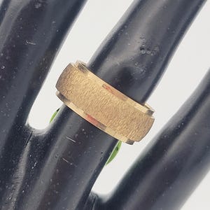 May include: A gold-colored ring with a textured band and a smooth, beveled edge. The ring is displayed on a black surface, showcasing its metallic sheen and design. The ring's band has a textured surface.