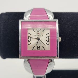 May include: A pink and silver square-shaped wristwatch with a white face. The watch face has black Roman numeral hour markers and a silver second hand. The watch face also has the text "DE NACCI QUARTZ JAPAN MOV'T".