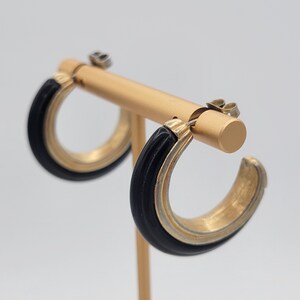 May include: A pair of gold-toned hoop earrings with a black, curved inlay. The earrings are displayed on a gold-colored earring stand against a white background. The earrings have a post-and-clasp closure.
