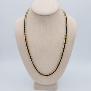 Puede incluir: Un collar dorado con un diseño de cuerda negra se exhibe en un maniquí beige. El collar es de estilo cuerda trenzada, con metal dorado y una cuerda negra entrelazada. El maniquí está sobre un fondo blanco.