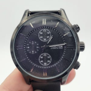 Puede incluir: Un reloj cronógrafo Kenneth Cole Reaction negro con una correa de malla negra. La esfera del reloj tiene tres subesferas y el nombre de la marca está impreso en la esfera.
