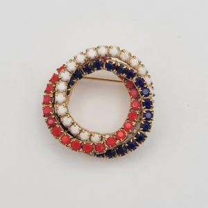 Puede incluir: Broche dorado con tres anillos entrelazados. Cada anillo está adornado con pequeñas piedras redondas rojas, blancas y azules. El broche tiene un cierre de alfiler. El diseño evoca los colores patrióticos.