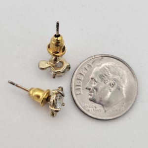 Puede incluir: Dos pendientes de bot&oacute;n dorados con piedras preciosas transparentes de corte cuadrado. Los pendientes est&aacute;n junto a una moneda para comparar el tama&ntilde;o. Los pendientes tienen un poste y un respaldo para un uso seguro.