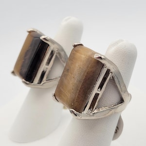 May include: Two rings with large, square tiger's eye stones set in silver-colored metal. The rings have a geometric design with triangular cutouts on the sides. The tiger's eye stones display bands of brown and gold.