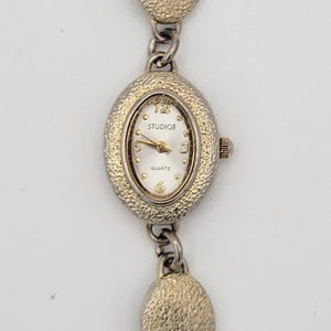 May include: A gold-toned oval-shaped wristwatch with a white face and black hands. The watch face has the text "STUDIO3" and "QUARTZ". The watch has a chain-link bracelet with oval-shaped links.