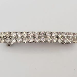 May include: A rectangular hair clip with two rows of clear, sparkling rhinestones. The clip has a silver-tone metal base and is designed to hold hair in place. The rhinestones are evenly spaced and catch the light, creating a glamorous look.