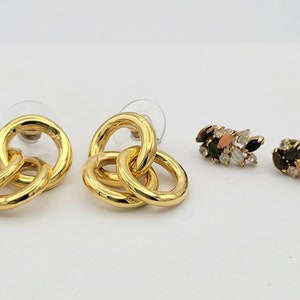 May include: Two pairs of gold earrings. One pair is a set of three interlocking rings. The other pair is a cluster of small, colorful stones.