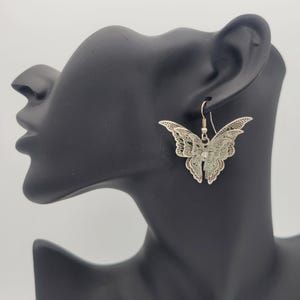 May include: Silver butterfly earrings with a delicate filigree design. The earrings are hanging from a mannequin's ear.