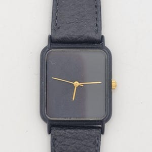 May include: A black wristwatch with a square face and a black leather band. The watch has gold-colored hands and a gold-colored crown. The watch band is approximately 8 inches long.