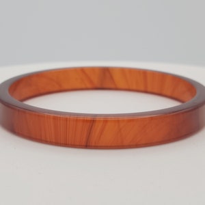 Puede incluir: Un brazalete redondo de color ámbar. El brazalete tiene una superficie lisa y pulida con un sutil patrón de vetas de madera. El brazalete mide aproximadamente 1,3 cm de ancho y es un accesorio de joyería clásico.
