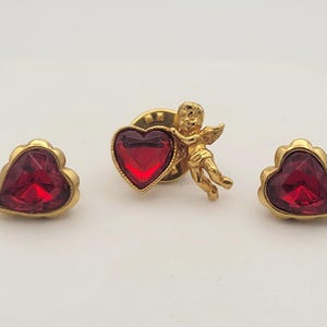Op de afbeelding: Een sieradenset met een goudkleurige cupidopspeld met een rood hart, geflankeerd door twee hartvormige oorstekers. De oorbellen en de speld hebben een gouden rand en een gefacetteerd rood midden.