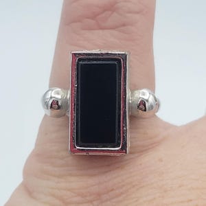 May include: Silver ring with a rectangular black stone center. The ring has a silver band with a red border around the black stone. The ring has two silver ball accents on either side of the rectangular stone.