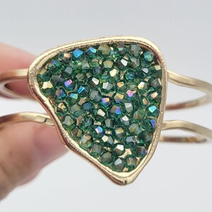 May include: Gold-tone bracelet featuring a unique design with a central, irregular-shaped element filled with sparkling green crystals. The bracelet has a double-band design for a modern look. A stylish piece of jewelry.