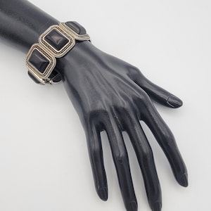 May include: A silver-toned bracelet with black rectangular stones. The bracelet is displayed on a black mannequin hand against a white background. The bracelet has a detailed design around the stones.