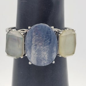 Puede incluir: Anillo de plata con tres piedras preciosas. La piedra central, ovalada, es azul, flanqueada por dos piedras rectangulares de color claro. El anillo se muestra sobre un soporte negro sobre un fondo blanco.