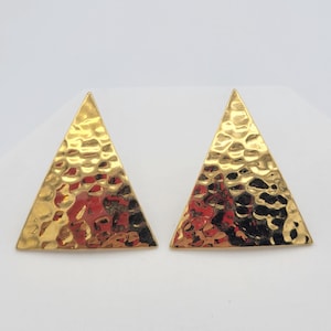 May include: A pair of gold-colored, hammered-texture triangle stud earrings. The earrings have a geometric design and a shiny finish. The earrings are displayed on a white background.