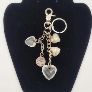 Puede incluir: Llavero plateado con múltiples dijes. Los dijes incluyen corazones de cristal transparente, corazones plateados texturizados y un pequeño dije en forma de bolso. El llavero tiene un cierre y un anillo para sujetar llaves.