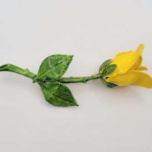 May include: A yellow rose brooch with green leaves and stem. The brooch is a detailed, enamel design, showcasing a realistic flower. The petals are a bright yellow, and the leaves are a deep green. The brooch is a classic floral accessory.