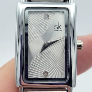 Könnte beinhalten: Eine rechteckige silberne Uhr mit weißem Zifferblatt und silbernem Mesh-Armband. Das Zifferblatt der Uhr hat ein diamantähnliches Design und der Text "sk SHENGKE" ist auf dem Zifferblatt aufgedruckt.