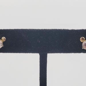 Puede incluir: Un par de pendientes de bot&oacute;n con un dise&ntilde;o dorado y un centro de piedra preciosa transparente y redondo. Los pendientes se exhiben en un expositor de joyer&iacute;a de terciopelo negro sobre un fondo blanco. Dise&ntilde;o inspirado en Mickey Mouse.