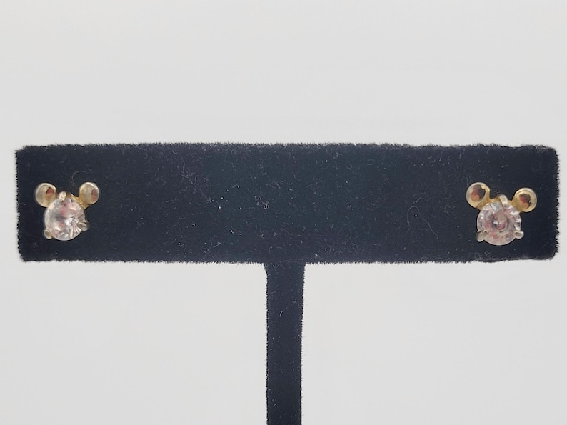 Puede incluir: Un par de pendientes de bot&oacute;n con un dise&ntilde;o dorado y un centro de piedra preciosa transparente y redondo. Los pendientes se exhiben en un expositor de joyer&iacute;a de terciopelo negro sobre un fondo blanco. Dise&ntilde;o inspirado en Mickey Mouse.