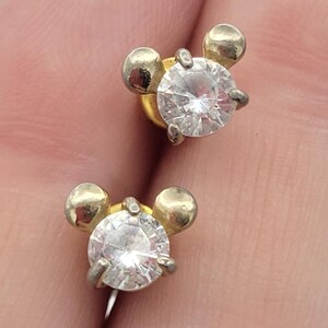 Puede incluir: Un par de pendientes de bot&oacute;n. Cada pendiente presenta una gema transparente de talla redonda engastada en metal plateado. El dise&ntilde;o incluye dos peque&ntilde;as esferas doradas a cada lado de la gema, creando una forma &uacute;nica.