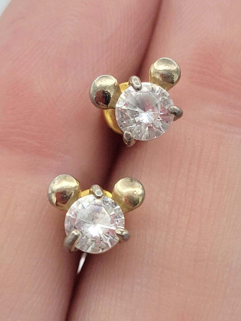 Puede incluir: Un par de pendientes de bot&oacute;n. Cada pendiente presenta una gema transparente de talla redonda engastada en metal plateado. El dise&ntilde;o incluye dos peque&ntilde;as esferas doradas a cada lado de la gema, creando una forma &uacute;nica.