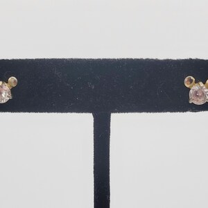 Puede incluir: Un par de pendientes de bot&oacute;n con una gema redonda y transparente en el centro y dos peque&ntilde;os acentos redondos a cada lado. Los pendientes se exhiben en un soporte negro de terciopelo sobre un fondo blanco.