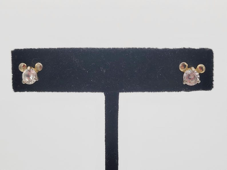 Puede incluir: Un par de pendientes de bot&oacute;n con una gema redonda y transparente en el centro y dos peque&ntilde;os acentos redondos a cada lado. Los pendientes se exhiben en un soporte negro de terciopelo sobre un fondo blanco.