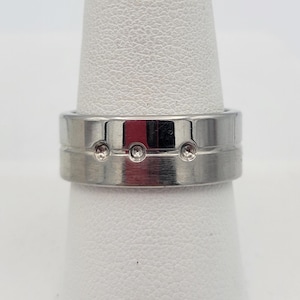 May include: A silver-colored ring with three small, circular embellishments. The ring has a band with a grooved design. The ring is shown on a white surface.