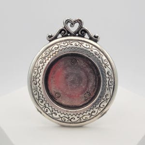Può includere: Un medaglione rotondo in metallo color argento con un bordo ornato da volute incise e un motivo a zigzag. La parte superiore del medaglione è decorata con un gancio a forma di cuore. Il centro del medaglione contiene un inserto circolare rosso.