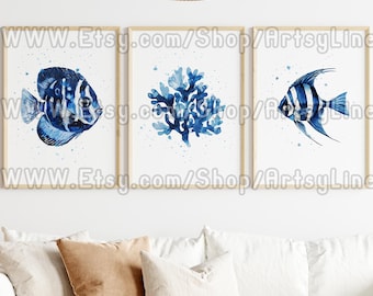 May include: Three framed watercolor prints featuring marine life. The prints showcase a blue fish, blue coral, and a striped blue and white fish. The frames are a light wood color, and the prints are displayed on a white wall above a white sofa.