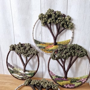 May include: Three circular wall hangings with woven trees and landscapes. Each piece features a brown tree with green leaves and a colorful woven landscape below. The trees are made of yarn and the landscapes are made of different colored yarns.
