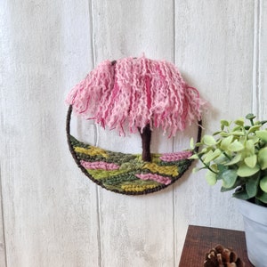 Half a 15cm hoop has been handwoveninthe bottom with a tree sculptured out of yarn ontheright hand side. The canopy of the tree is made up of yarn dangling from the top of the hoop in varying shades of pink.