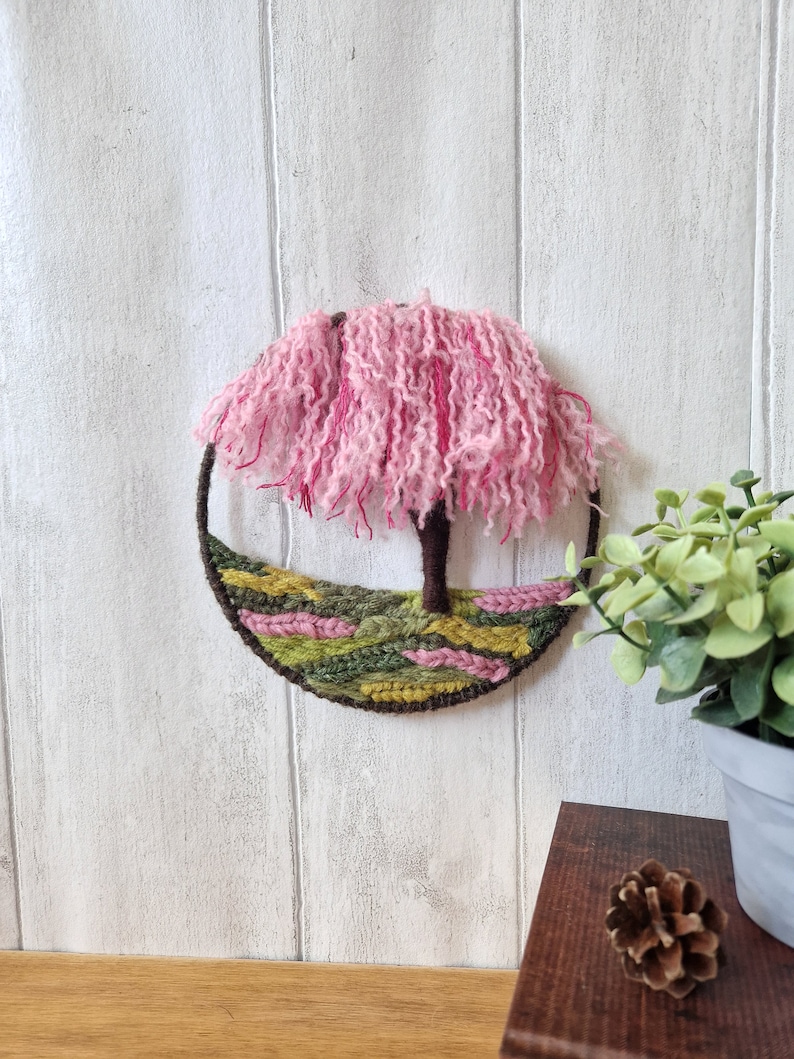 Half a 15cm hoop has been handwoveninthe bottom with a tree sculptured out of yarn ontheright hand side. The canopy of the tree is made up of yarn dangling from the top of the hoop in varying shades of pink.