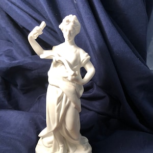 May include: A white porcelain figurine of a woman in a flowing robe, holding a scroll in one hand and a torch in the other. The figurine is standing on a base that resembles a rock.