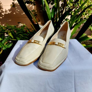 May include: Pair of off-white leather loafers with gold buckles. The loafers have a perforated design and a square toe.