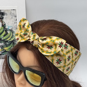 Puede incluir: Una diadema estampada con lazo, con fondo crema y diseño geométrico en amarillo, verde y rojo. La diadema se lleva en una cabeza de maniquí con gafas de sol oscuras.
