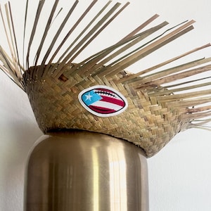 Puede incluir: Un sombrero de paja tejido con un parche decorativo con la bandera de Puerto Rico en forma de balón de fútbol. El sombrero está colocado sobre una base metálica dorada y redondeada. El sombrero tiene un diseño de rayos de sol.