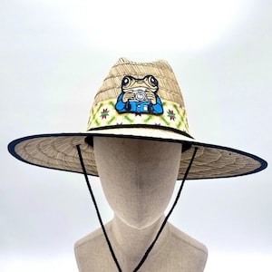 Puede incluir: Sombrero de paja con ala ancha y correa de barbilla negra. El sombrero presenta una banda colorida con un patrón repetitivo y un parche de una rana sosteniendo una cámara. El ala tiene un ribete azul oscuro.