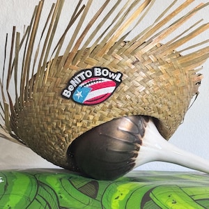 Puede incluir: Un sombrero de paja tejido con un parche decorativo que dice "Benito Bowl" y presenta la bandera de Puerto Rico. El sombrero se combina con un objeto de cerámica blanco y marrón, que descansa sobre una superficie verde.
