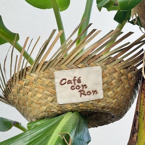Puede incluir: Cesta tejida con un parche rectangular que dice "Café con Ron" en bordado marrón. La cesta está hecha de material tejido marrón claro y verde. La cesta está rodeada de hojas y tallos verdes.