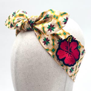 Puede incluir: Una diadema color crema con un estampado geométrico en amarillo, verde y burdeos. Una gran flor de hibisco roja bordada con detalles negros y amarillos está adherida. La diadema está atada con un lazo.