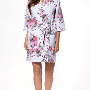 May include: White waffle-weave robe with a floral pattern in shades of pink, purple, and green. The robe has a V-neck, three-quarter sleeves, a matching belt, and two front pockets. The robe is knee-length.